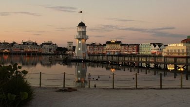Disney's BoardWalk lighthouse and pier shine on the lagoon at sunset, with resort buildings and Atlantic Dance Hall in view.