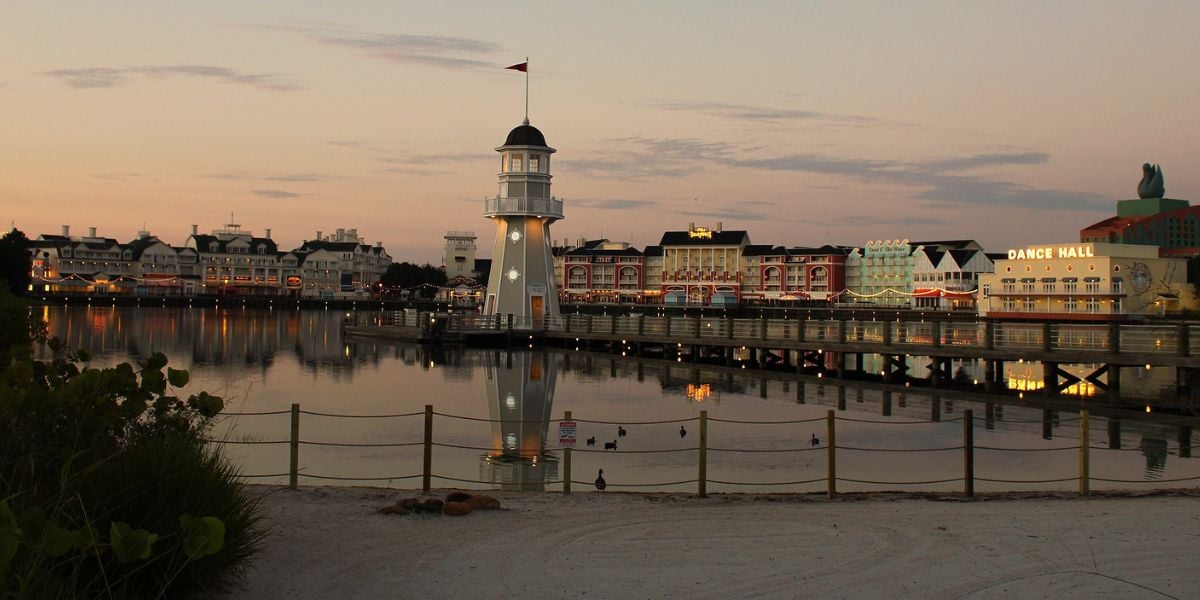 Disney's BoardWalk lighthouse and pier shine on the lagoon at sunset, with resort buildings and Atlantic Dance Hall in view.