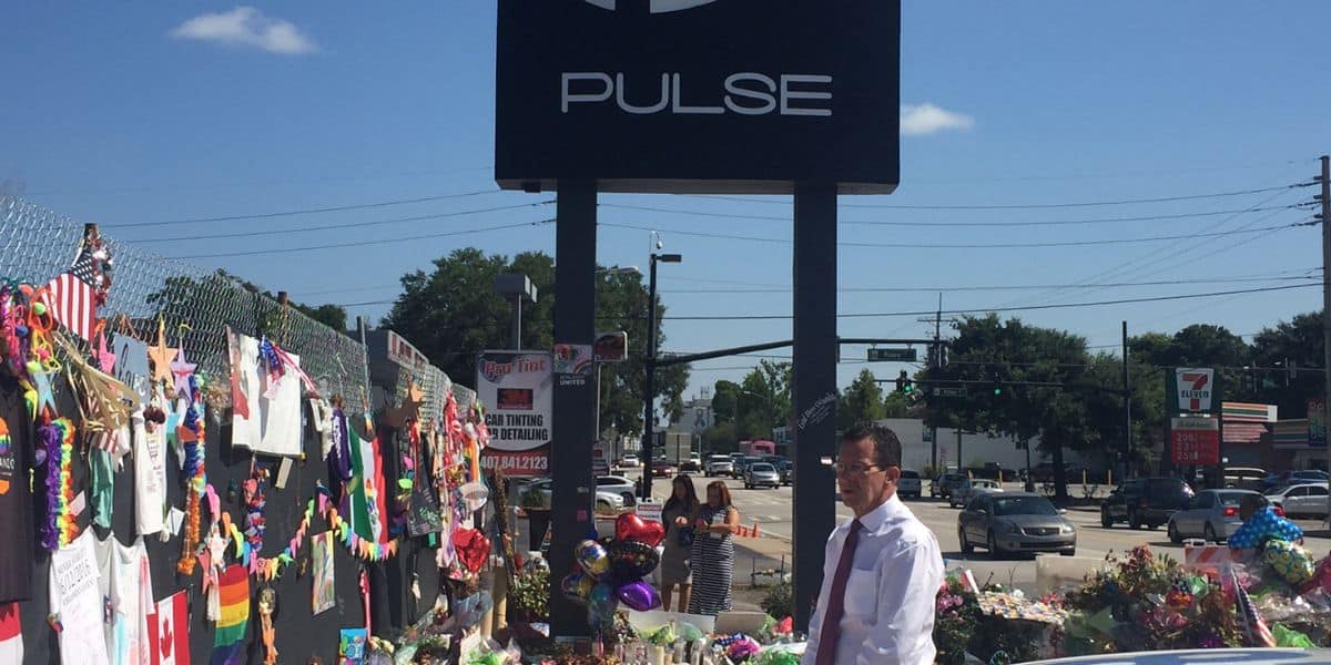 A guest pauses by a tribute outside Pulse nightclub, where flowers and messages honor victims beneath the iconic Pulse sign.