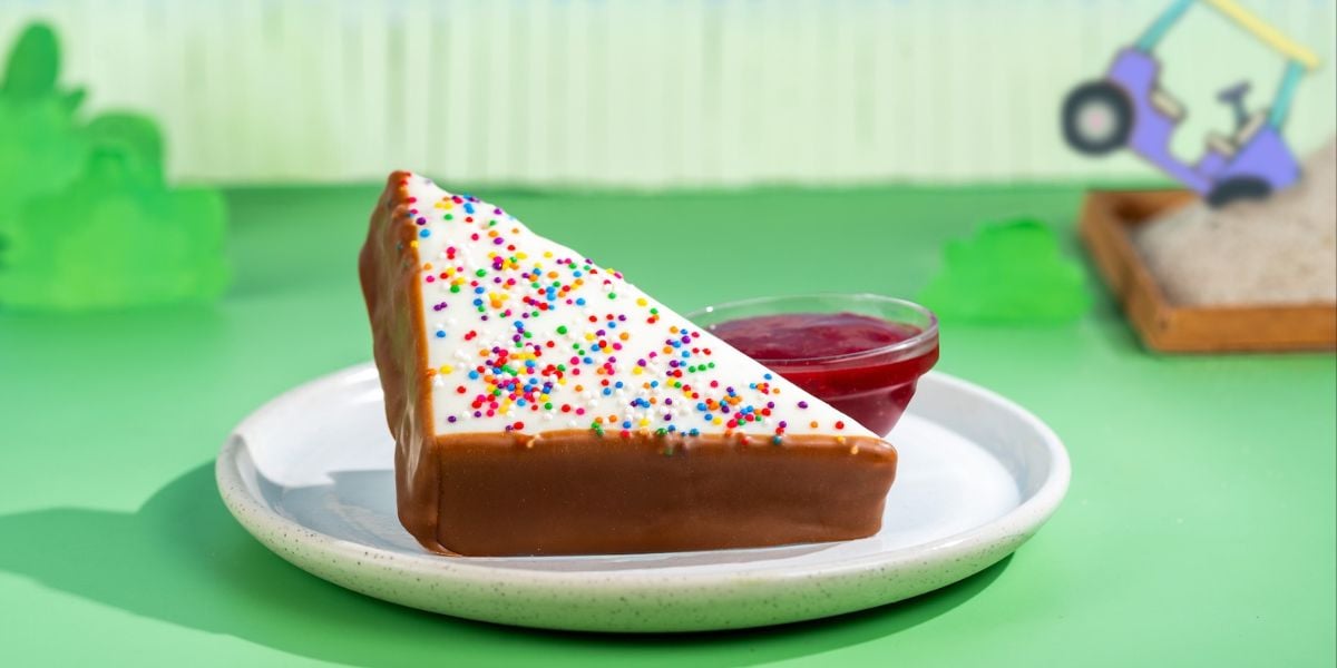 A festive slice of sprinkle-topped cake with white icing and red sauce, served on a plate at the heart of theme park fun.