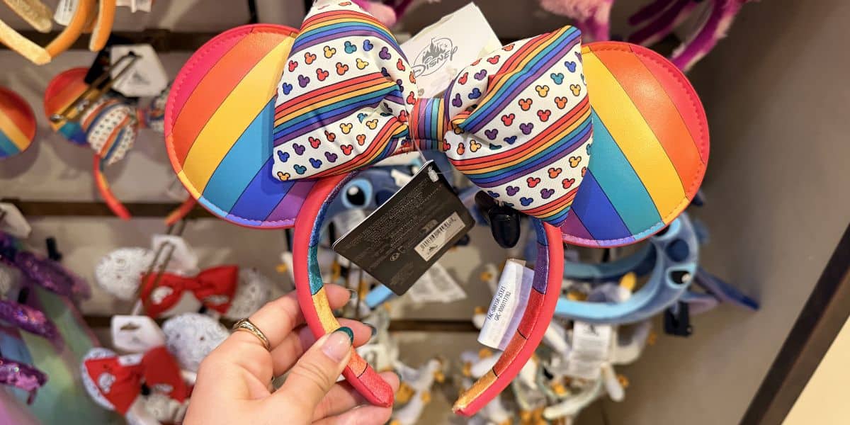 Rainbow-striped Minnie Mouse ears with a colorful heart bow, perfect for Pride Month celebrations and LGBTQ+ support at Disney Parks.