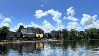 Disney Orlando Resort’s new 2027 hotel reflects in a lake, framed by trees and blue skies; booking limits may apply.