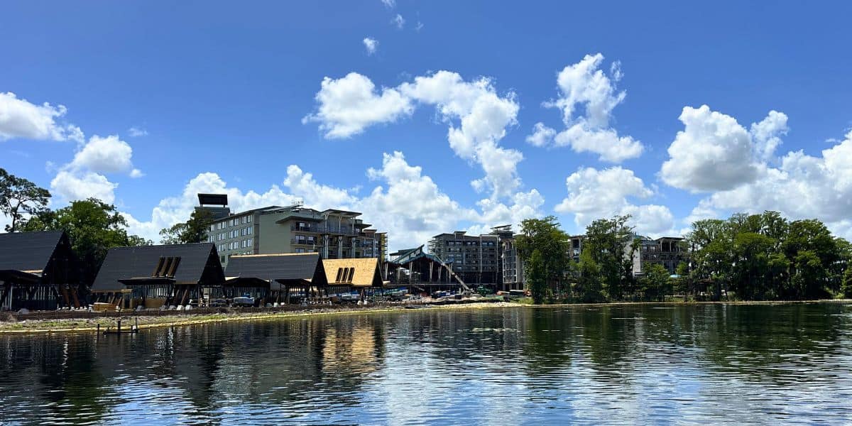 Disney Orlando Resort’s new 2027 hotel reflects in a lake, framed by trees and blue skies; booking limits may apply.