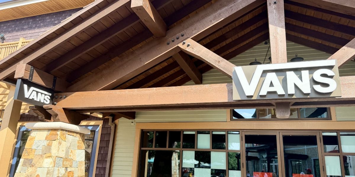 Rustic storefront at a lively shopping district, featuring two bold Vans signs—one above the entrance and one on a stone pillar.