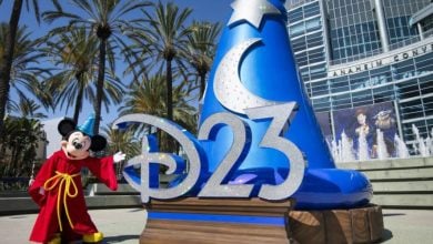 Mickey Mouse, dressed in his signature red robe, greets guests beside the dazzling blue sorcerer hat and bold "D23" sign outside the Anaheim convention center, setting a magical tone for fans entering the lively show floor.