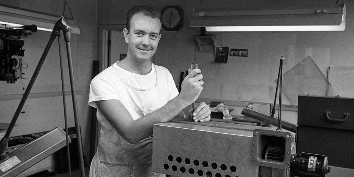 An Imagineer grins in his workshop, showcasing a tool beside an impressive piece of machinery—radiating the inventive spirit behind classic Disney magic like that seen in Mary Poppins.