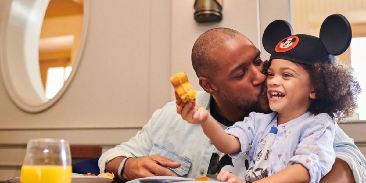 A dad shares a sweet moment with his smiling child wearing Mickey Mouse ears, who’s enjoying a fresh waffle at Disney World; a glass of orange juice sits on the table.