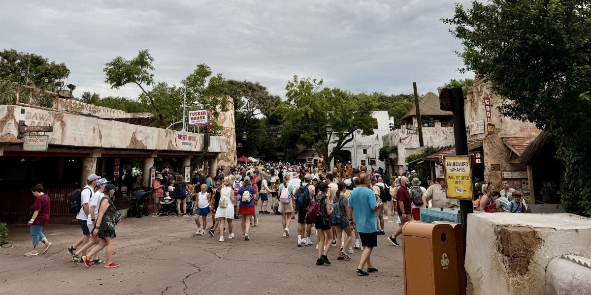Guests explore a bustling section of the theme park, strolling past charming, rustic buildings nestled among lush trees beneath an overcast sky.