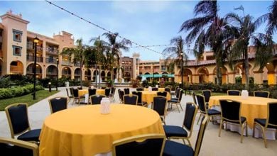 An inviting outdoor courtyard at Walt Disney World Resort, featuring round tables draped in cheerful yellow cloths, twinkling string lights overhead, and swaying palm trees—a perfect spot for guests to unwind and find comfort after the recent park closure.