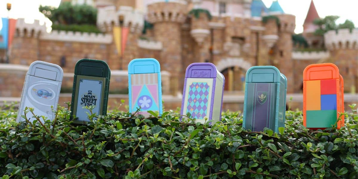 Six colorful miniature trash cans are arranged atop a well-manicured hedge in front of a grand, castle-inspired building, capturing the playful and detailed charm that fans love about Magic Kingdom and Disney’s latest park experiences.