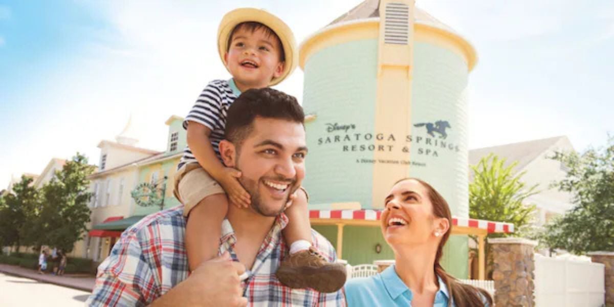 Happy family outside Disney’s Saratoga Springs Resort & Spa, child riding on dad’s shoulders, enjoying the excitement and special experiences that come with a Disney resort stay.