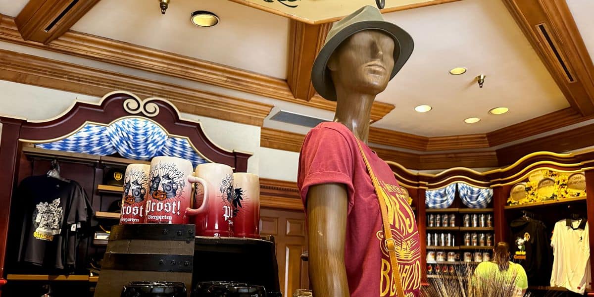 The Germany Pavilion at EPCOT invites guests to browse a cozy gift shop, where a mannequin dressed in a red shirt and hat showcases themed merchandise and mugs amid rustic wood decor.