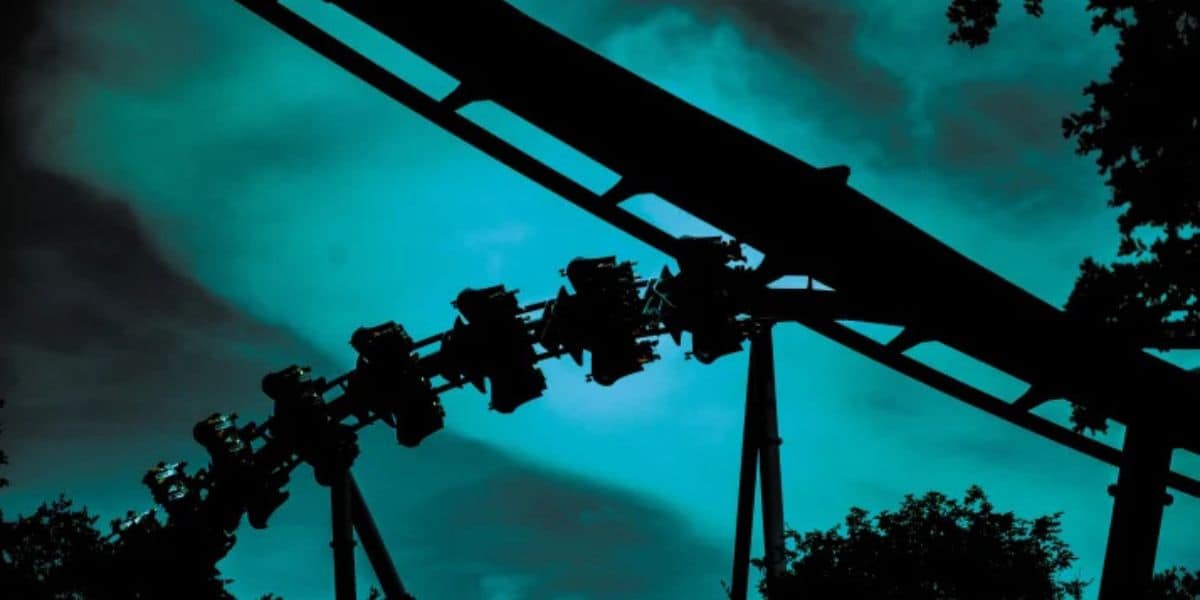 Riders soar on a thrilling roller coaster at the theme park, silhouetted against a dramatic teal sky with looming dark clouds.