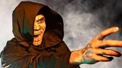 A ghoulish character from the park’s haunted attraction, cloaked in a hooded robe, reaches out with a pale-painted face and open mouth, enveloped by swirling smoke effects.