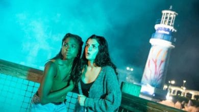Two guests huddle together on the boardwalk at night in front of a brightly illuminated lighthouse, their expressions filled with suspense and excitement, capturing the thrill of a theme park adventure after dark.