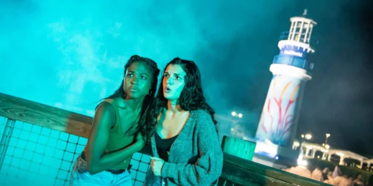 Two guests huddle together on the boardwalk at night in front of a brightly illuminated lighthouse, their expressions filled with suspense and excitement, capturing the thrill of a theme park adventure after dark.