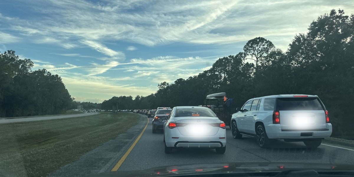 Guests’ cars are lined up in bumper-to-bumper traffic at a theme park exit during the day, with lush park trees and whimsical cloud-shaped landscaping visible in the background.