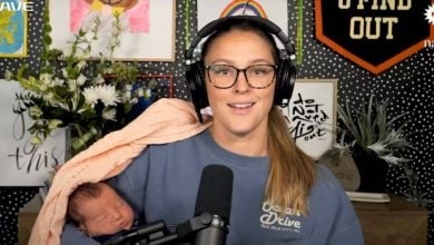 A podcast host with headphones broadcasts live, cradling her peacefully napping baby in a pink wrap, backstage at Universal Studios.
