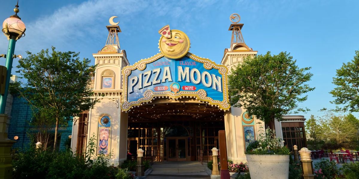 Pizza Moon at Epic Universe stands out with its whimsical, smiling moon sign, surrounded by lush trees and a clear sky. This inviting restaurant promises theme park guests an unforgettable pizza experience that captures the cosmic magic of the park right at your table.