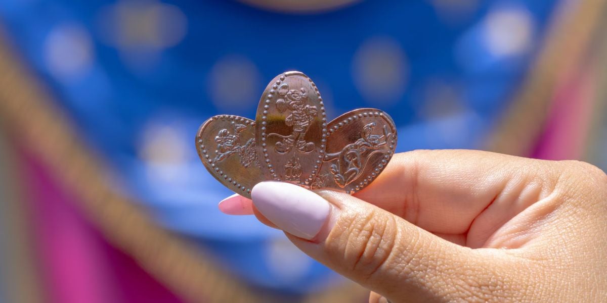 A guest with pink nails shows off three shiny pressed souvenir coins, captured against the vibrant blue and pink lights of the park.
