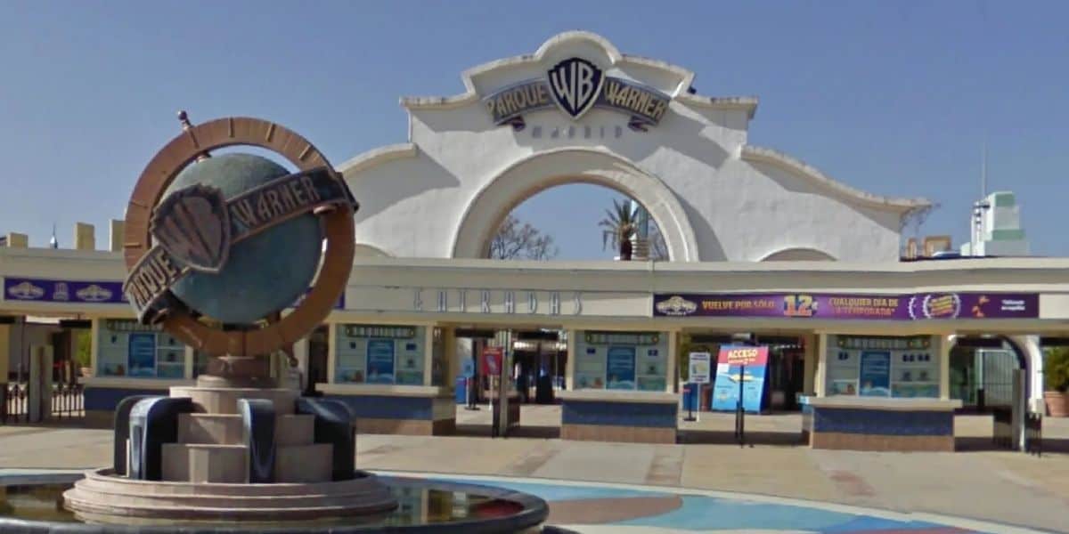 Parque Warner Madrid entrance featuring the iconic WB shield statue and colorful ticket booths, all welcoming guests under a bright blue sky.
