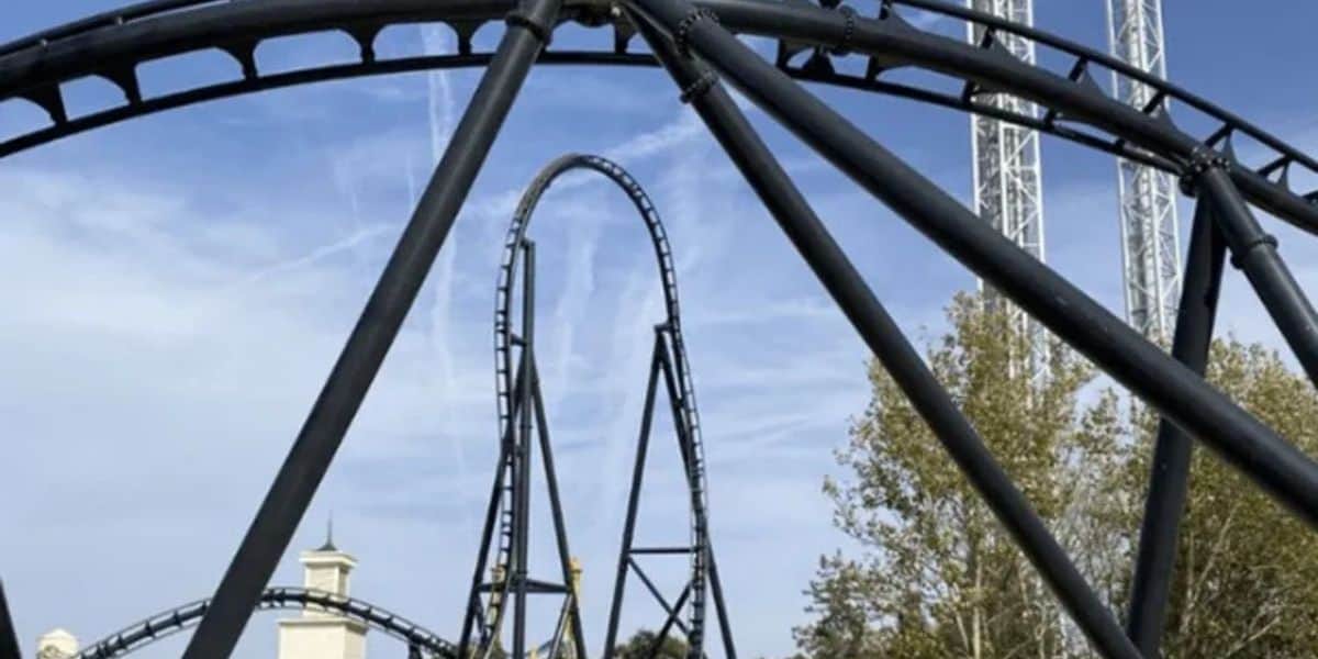 Twisting black steel coaster track packed with thrilling loops set against a bright blue sky, surrounded by lush trees and towering white support structures at the amusement park.