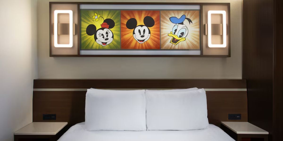 A Disney World Resort hotel room features a bed beneath whimsical artwork of Mickey Mouse and Donald Duck, framed by two wall lamps. This playful decor hints at the immersive experience awaiting fans when the new resort opens in 2027.