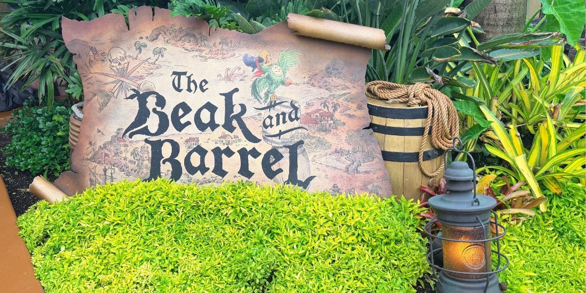 The weathered “The Beak and Barrel” sign welcomes guests at the entrance, surrounded by lush greenery, a classic barrel, and an old-fashioned lantern—inviting visitors to experience the cozy tavern’s themed seating area just one year into its storybook adventure.