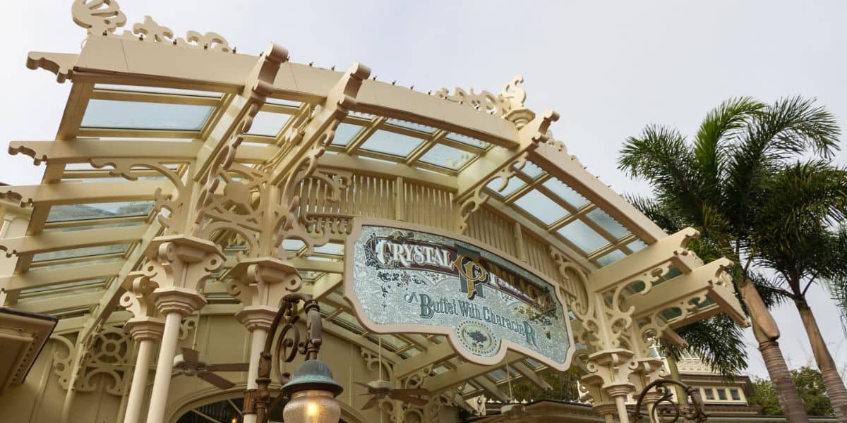 Elegant glass and iron entrance of the Crystal Palace restaurant at Magic Kingdom, with a tropical palm tree and Victorian-style lantern enhancing the enchanting theme park atmosphere.