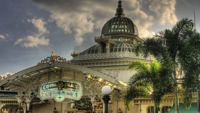 The iconic Crystal Palace with its intricate glass dome rises above lush palm trees, set against a backdrop of dramatic clouds—an inviting landmark at the heart of the park, promising guests a grand Victorian-themed dining and entertainment experience.