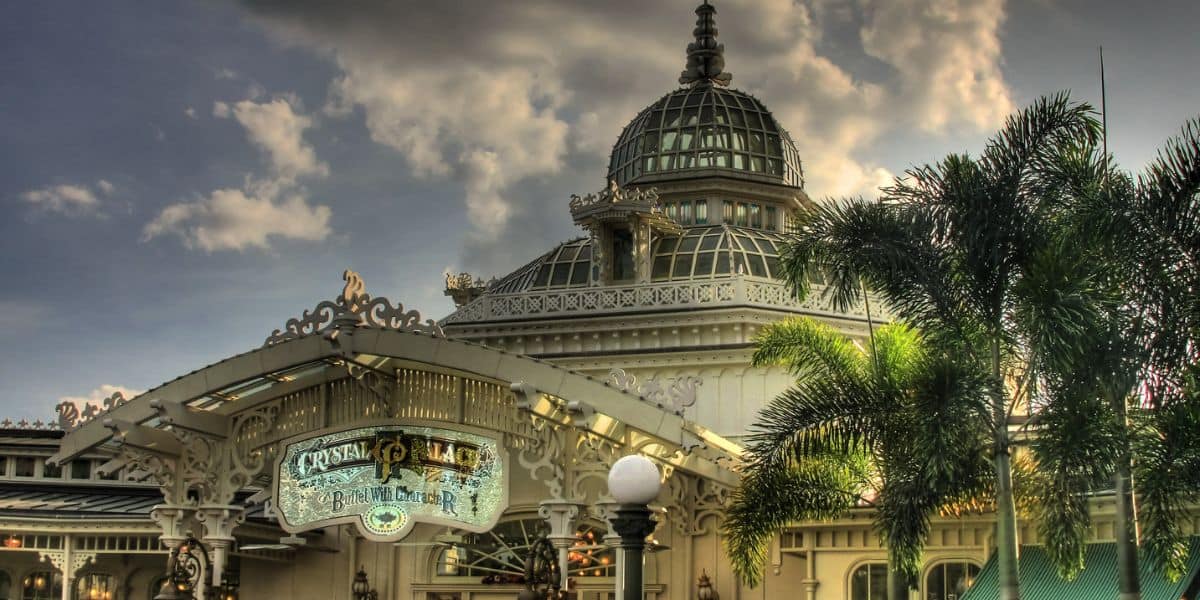 The iconic Crystal Palace with its intricate glass dome rises above lush palm trees, set against a backdrop of dramatic clouds—an inviting landmark at the heart of the park, promising guests a grand Victorian-themed dining and entertainment experience.