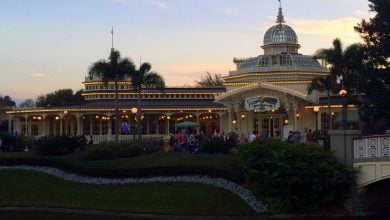 The Crystal Palace at Magic Kingdom glowing with string lights at sunset, nestled among lush gardens and swaying palm trees.
