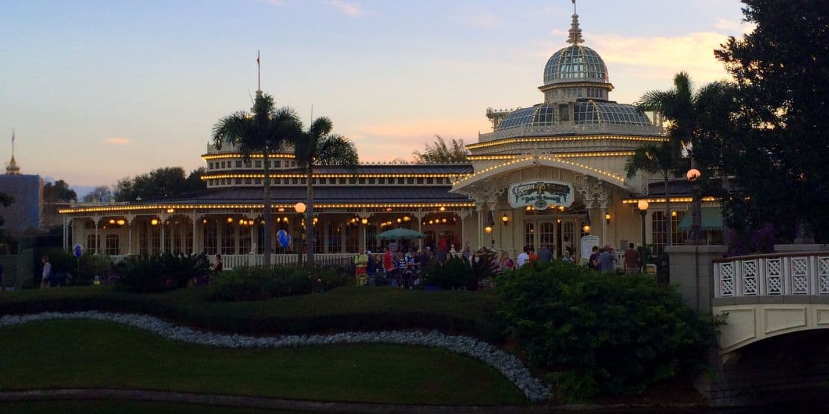 The Crystal Palace at Magic Kingdom glowing with string lights at sunset, nestled among lush gardens and swaying palm trees.