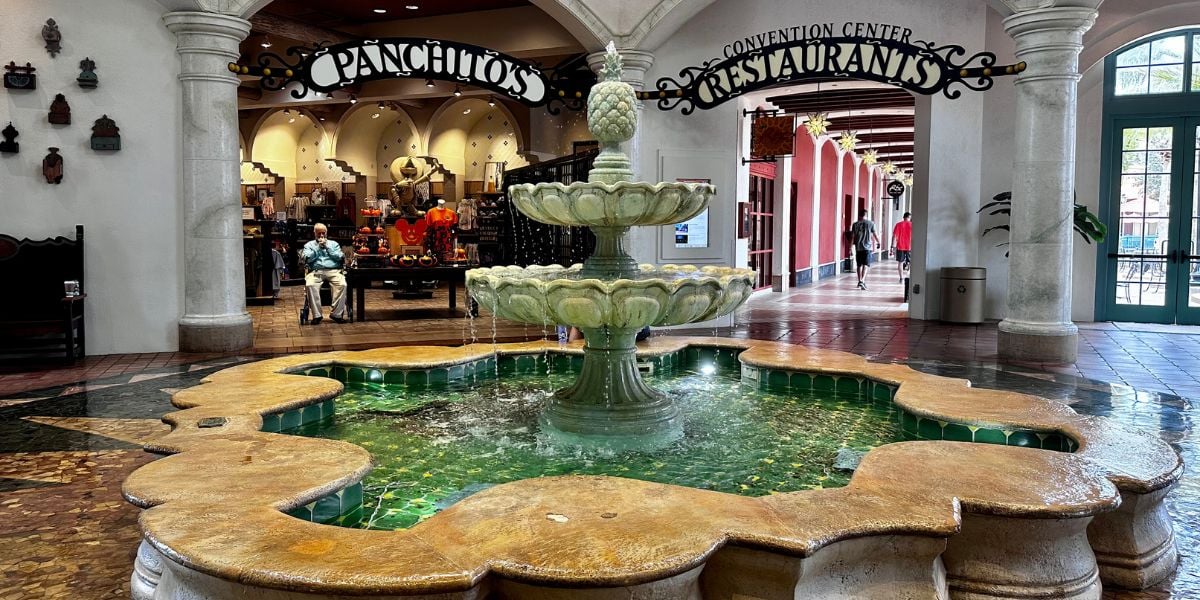A beautifully tiled stone fountain with vibrant green accents stands inside a Disney World pavilion, surrounded by nearby shops and a welcoming sign pointing guests to popular restaurants.
