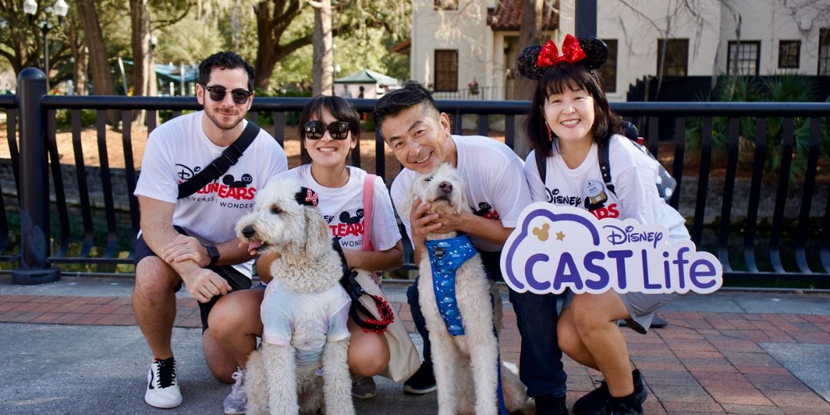 A group of four Disney Cast Members, all grinning and wearing classic Mickey ears, join two cheerful poodles for a photo as they proudly hold up a “Disney CAST Life” sign. The image celebrates the spirit of Disney wellness and camaraderie among Cast Members in the magical world of Disney Parks.