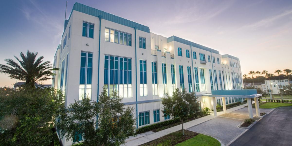 A sleek white and blue Disney building with expansive windows stands amid lush trees, set against a glowing sunset sky, welcoming both guests and Cast Members into a space inspired by Disney’s commitment to wellness, comfort, and magical hospitality.