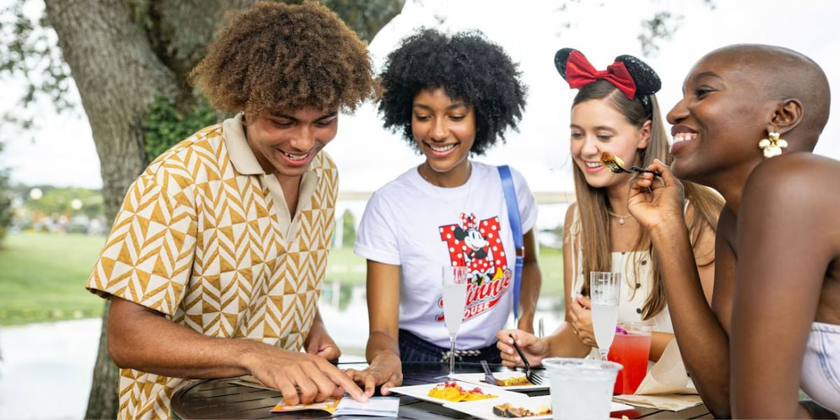 Guests gather at an EPCOT Food & Wine Festival outdoor table, smiling as they browse festival menus and indulge in signature dishes and drinks amidst the park’s lively atmosphere.