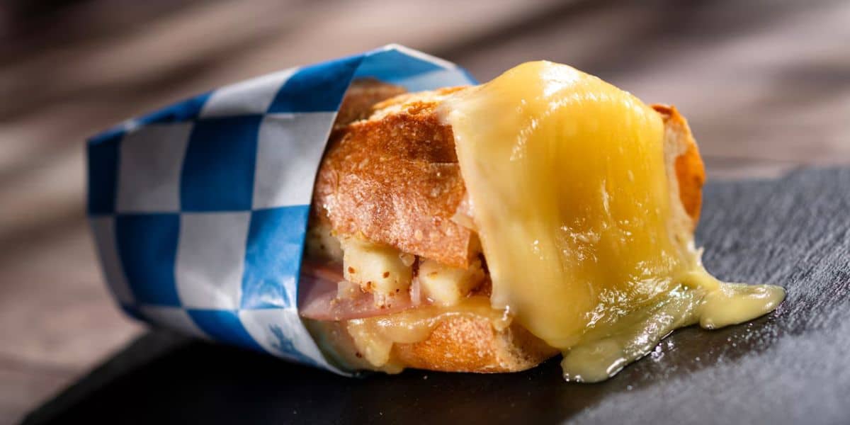 A mouthwatering baguette sandwich in signature blue EPCOT Food & Wine Festival wrap, with gooey melted cheese spilling out onto a dark table—just like the cheesy delights you’d savor at World Showcase.