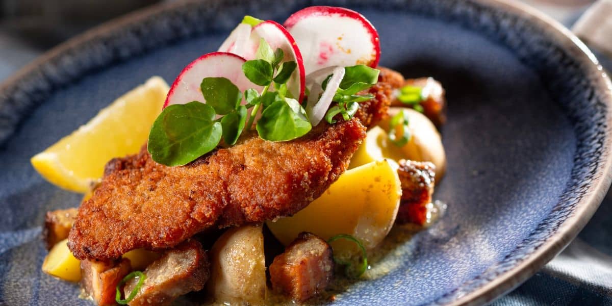 A crispy breaded cutlet served with golden roasted potatoes, fresh lemon wedges, radish slices, and greens on a vibrant blue plate—bringing the magic of EPCOT Food & Wine Festival 2026 to Disney World fans through this delicious festival-inspired dish.