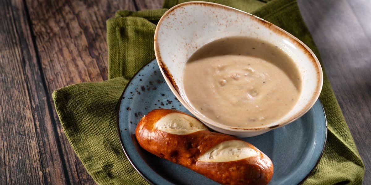 A bowl of creamy soup with a side of pretzel bread on a plate, set atop a green napkin on a wooden table—this EPCOT Food & Wine Festival-inspired dish is perfect for Disney fans eager to savor an iconic taste while counting down to the excitement of the 2026 Disney World Festival.