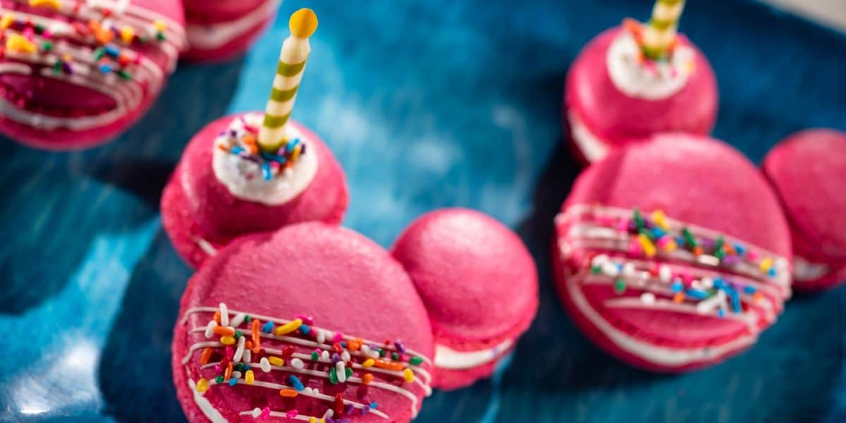 Bright pink macarons with white filling and playful sprinkles, some crowned with striped birthday candles, are displayed on a blue plate—an Instagram-ready treat sure to delight Disney World fans at the 2026 Festival or EPCOT Food & Wine event.