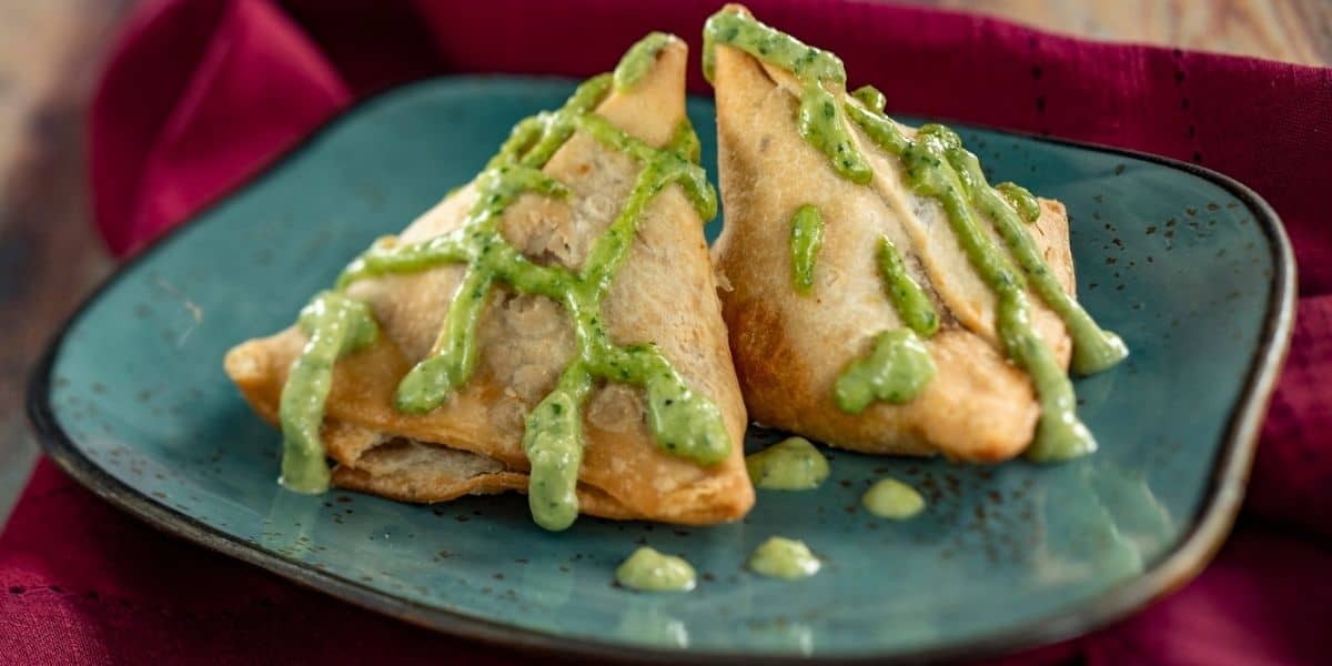 A pair of crispy golden samosas drizzled with vibrant green chutney, served on a blue plate over maroon tablecloth at the colorful Festival of Flavors food booth in Adventure Park.