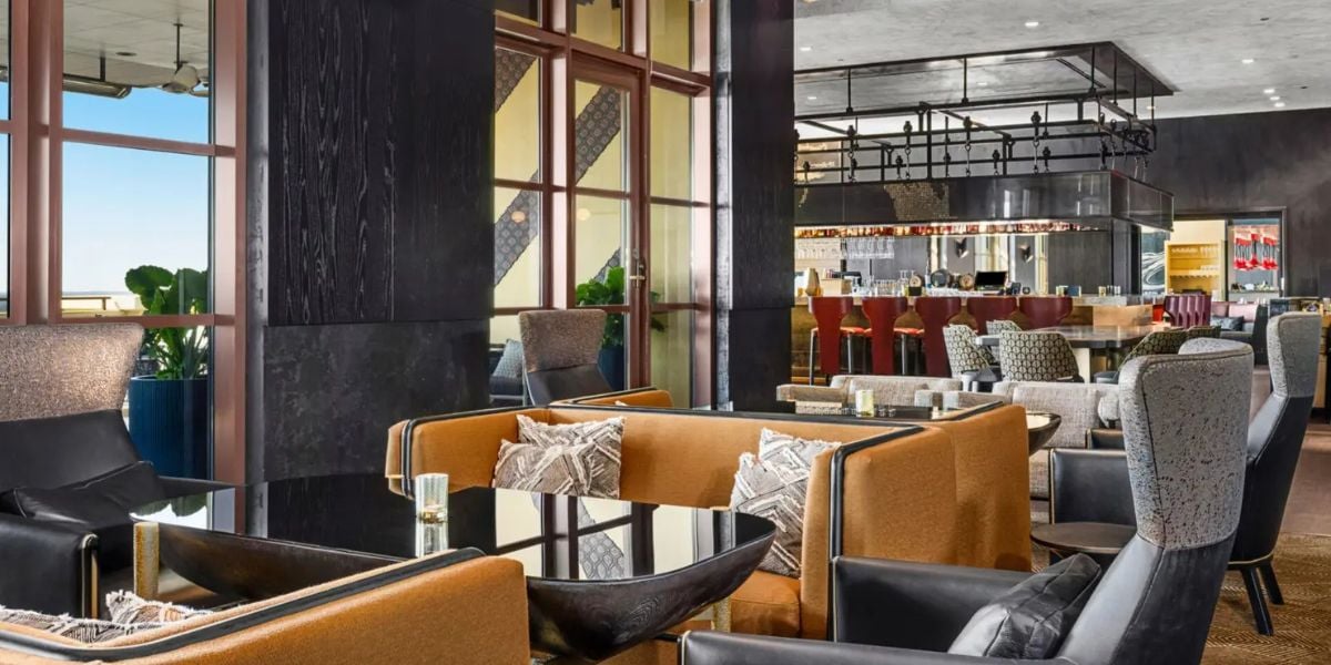 Upscale lounge area at a theme park resort, featuring comfy seating, sleek glass tables, panoramic windows overlooking the attractions, and a vibrant bar where guests can relax between roller coasters.