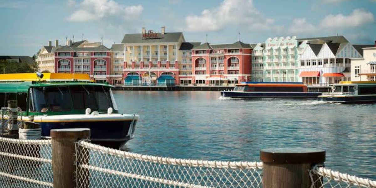 Vibrant Disney resort buildings and charming boats line the scenic waterfront, with a rope fence in the foreground and clear blue skies overhead—a lively, immersive atmosphere perfect for theme park fans soaking up exclusive Disney resort benefits.
