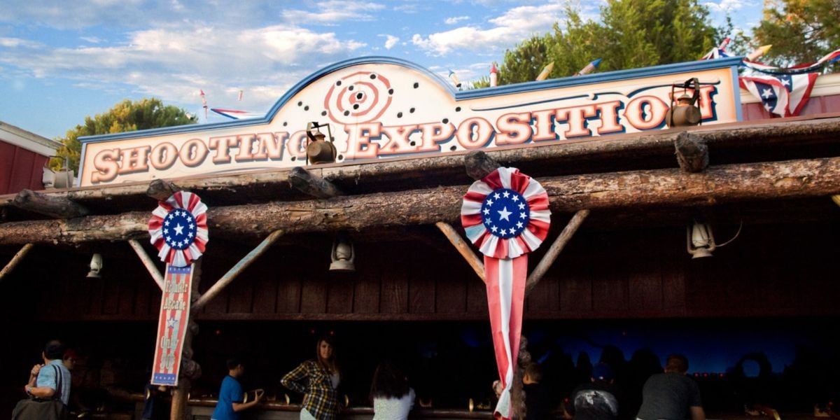 Shooting Exposition entrance decked out in red, white, and blue decorations, excited guests gathering below, and lush trees surrounding the area beneath a bright blue sky.