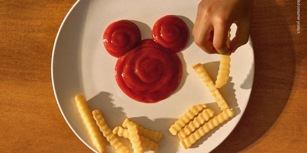 Crinkle fries on a plate spell out “Hey” next to ketchup arranged in the iconic shape of Mickey Mouse’s head—a cheerful tribute to Disneyland Resort magic. A hand picks up one fry, emphasizing the whimsical spirit and tasty fun brought by this Kraft Heinz collaboration for Disney fans.