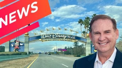 Disney World President smiles in a sharp suit at the entrance, standing beside a bold “NEWS” banner as he shares thrilling updates ahead of his big homecoming celebration.