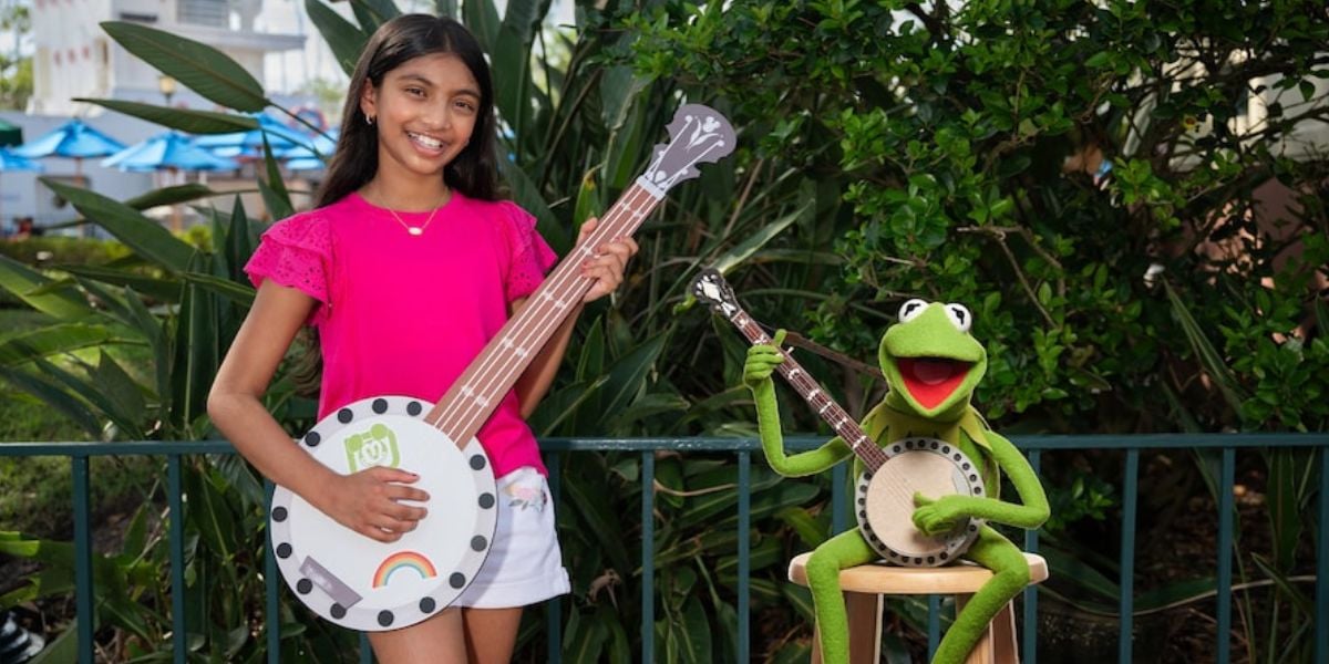 Kermit the Frog and a young guest strum banjos together in an outdoor setting surrounded by lush greenery, bringing classic Muppet fun to life for visitors.