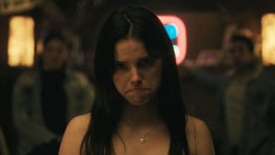 A woman with long dark hair frowns in moody, shadowy lighting inside a room, while two out-of-focus figures linger behind her, channeling the chilling atmosphere of Halloween Horror Nights at Universal Studios.