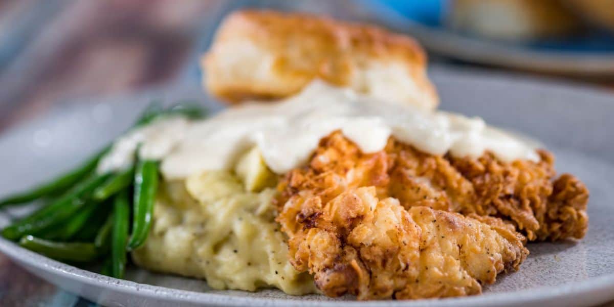 A plate piled high with crispy fried chicken, creamy mashed potatoes topped with savory gravy, fresh green beans, and a fluffy biscuit captures the delightful flavors of Disney World dining—bringing the joy and nostalgia of your favorite Magic Kingdom meal to every bite.